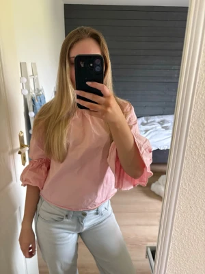 Ljusrosa blus med volangärmar - Söt ljusrosa blus med vida trekvartsärmar och volangdetaljer. Blusen har en lös passform och knytning i nacken med en liten öppning baktill. Perfekt för en trendig och avslappnad look. Använd en gång