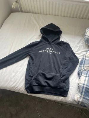Svart hoodie Peak Performance - Snygg svart hoodie från Peak Performance med vit logga på bröstet. Klassisk modell med huva och stor magficka framtill. Tillverkad i mjukt bomullsmaterial som är skönt att ha på sig. Perfekt för dig som gillar en clean och sportig stil.