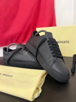Svarta sneakers från Axel Arigato - Stilrena svarta sneakers från Axel Arigato i slät läderfinish. Klassisk låg modell med svarta skosnören och diskret guldfärgad logga på sidan. Perfekta för dig som gillar minimalistisk och clean stil.