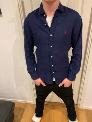 Mörkblå skjorta från Ralph Lauren - Klassisk mörkblå skjorta från Ralph Lauren med vit knappstängning och röd broderad logga på bröstet. Skjortan har lång ärm och normal passform, tillverkad i mjuk bomull. Perfekt för en stilren och avslappnad look.