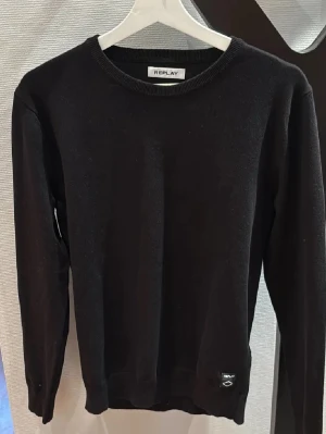  Replay - Klassisk svart stickad tröja från Replay med rund halsringning och diskret logga nertill. Tröjan har lång ärm och ribbade muddar vid ärmslut och nederkant. Perfekt basplagg för lager på lager och enkel att matcha.