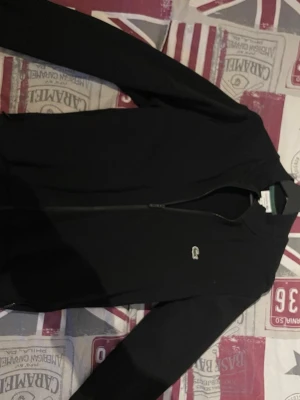 Svart ziptröja från Lacoste - Snygg svart ziptröja från Lacoste med klassisk krokodillogga på bröstet. Tröjan har hel dragkedja framtill, ribbade muddar och en enkel, stilren design som passar till det mesta. Perfekt för dig som gillar sportig och clean stil. Storlek S.