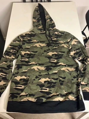Camo hoodie med svart kant - Säljer en camouflagemönstrad hoodie från WN med klassisk huva och svarta detaljer. Hoodien har en stor magficka och ribbad svart kant nedtill. Perfekt för dig som gillar streetwear och vill sticka ut med en snygg camo-look.