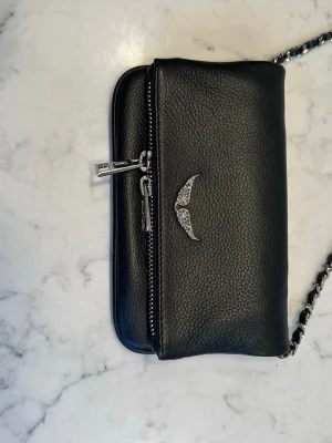 Svart handväska med kedjerem Zadig & Voltaire - Säljer en svart handväska i skinnimitation med silverfärgad kedjerem och dragkedja. Väskan har en snygg detalj med två silverfärgade vingar på framsidan. Perfekt för dig som gillar stilrena accessoarer med en twist.