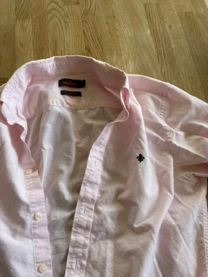 Ljusrosa skjorta från Morris - Säljer en ljusrosa skjorta från Morris med klassisk krage och lång ärm. Skjortan har knappar framtill och en broderad logga på bröstet. Tillverkad i mjuk bomull som känns skön mot huden. Perfekt för en stilren och avslappnad look. Köpt på Vinted men var tyvärr för stor så säljs vidare. Storlek S- 37/38