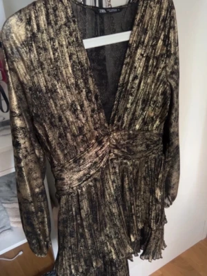  kort klänning från Zara - Kort glittrig klänning från Zara i svart och guld med djup v-ringning, långa ärmar och markerad midja. Kjolen är plisserad och tyget har ett skimrande metallicmönster som verkligen syns. Perfekt för dig som vill ha en statementklänning.