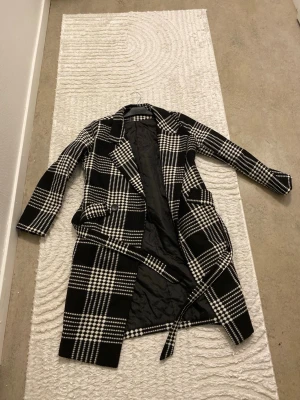 Svartvit rutig kappa med bälte - Stilren svartvit rutig kappa med bälte i midjan och stora slag. Kappan har två framfickor och är helfodrad med svart innerfoder. Perfekt för dig som vill ha en trendig och klassisk look. Passar till många olika outfits.