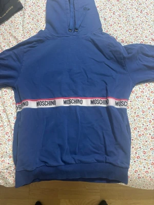 Blå hoodie från Moschino - Snygg blå hoodie från Moschino med huva och dragsko. Hoodien har vita och rosa band med Moschino-loggan runt midjan och ärmarna, samt ribbade muddar vid ärmslut och nederkant. Perfekt för dig som gillar streetwear och vill sticka ut.