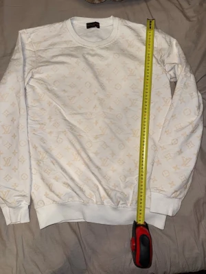Vit Louis Vuitton sweatshirt - Säljer en vit sweatshirt från Louis Vuitton med diskret beige monogrammönster över hela tröjan. Tröjan har rund halsringning, långa ärmar och ribbade muddar vid ärmslut och nederkant. Perfekt för dig som gillar exklusiv streetwear.