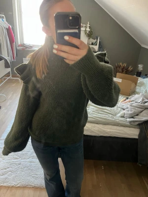 Grön stickad tröja med volanger Vila - Mysig mörkgrön stickad tröja från Vila med volangdetaljer på ärmarna och hög hals. Tröjan har en avslappnad passform och långa ärmar, perfekt för att hålla värmen under kyliga dagar. Materialet är mjukt och ger en skön känsla.