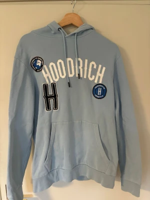 Ljusblå hoodie från Hoodrich - Snygg ljusblå hoodie från Hoodrich med vita och svarta tryck och patches på bröstet. Stor magficka framtill, dragsko i huvan och ribbade muddar. Hoodien har en avslappnad passform och är tillverkad i mjukt bomullsmaterial. Perfekt för en chill och street-inspirerad look.