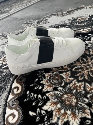 Vita Valentino sneakers med svart detalj - Snygga vita sneakers från Valentino med bred svart rem över vristen och coola nitar på hälen. Skorna har vita snören, låg profil och en chunky sula med rutmönster undertill. Perfekta för dig som vill ha en stilren men edgy look. VI KAN DISKUTERA OM PRISET!!!!!!!!!!!!!!