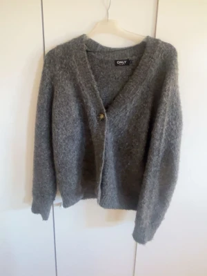 Grå fluffig kofta från ONLY - Mysig grå kofta från ONLY med fluffig textur och v-ringning. Koftan har en knapp framtill och långa ärmar. Perfekt för lager på lager och ger en avslappnad men ändå stilren look.