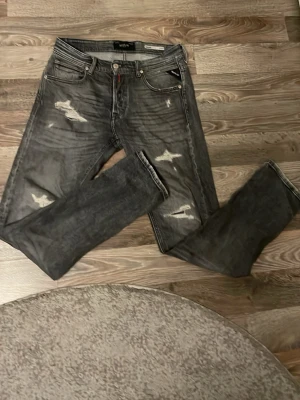 Grå slitna jeans från replay - Säljer ett par grå jeans från replay med slitningar och distressed detaljer på både fram- och baksida. Klassisk femficksmodell med raka ben och knappgylf. Perfekta för en avslappnad och trendig look.