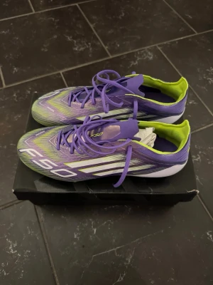 Adidas F50 lila och grön fotbollsskor - Säljer ett par Adidas F50 fotbollsskor i en riktigt snygg lila och limegrön färgkombination. Skorna har klassiska snörningar, låg profil och mönstrad ovandel för bra bollkontroll. Yttersulan har fasta dobbar i lila och lime, perfekt för spel på gräsplan.