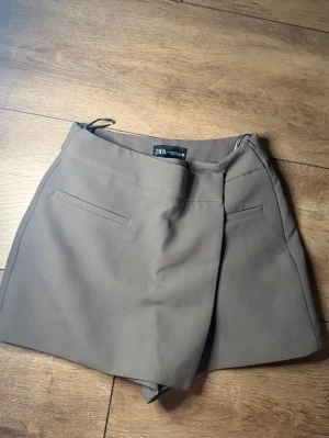 Beige skort från Zara - Stilren beige skort från Zara med snygg omlott-look framtill och två dekorativa fickor bak. Skorten har en klassisk passform och är gjord i ett mjukt, vävt material som känns bekvämt hela dagen. Perfekt för dig som gillar en clean och trendig stil.