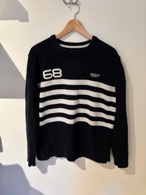 Svart stickad tröja Morris - Svart stickad tröja från Morris med vita horisontella ränder över bröstet och ryggen. Siffran 68 och Morris Company-logga på bröstet samt Union Jack-detalj på ärmen. Rund hals och lång ärm. Perfekt för en sportig och stilren look.