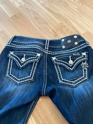 Y2K Miss Me mörkblå jeans med detaljer - Säljer ett par helt nya mörkblå jeans från Miss Me med kontrastsömmar och snygga bakfickor med dekorativa knappar och broderade detaljer. Jeansen har en klassisk femficksmodell och coola slitningar för en trendig look. Storlek 27!!
