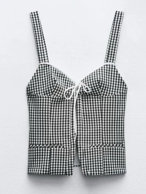 Rutig korsettopp med snörning - Snygg svartvit rutig korsettopp med breda axelband och söt snörningsdetalj framtill. Toppen har figurnära passform och markerade sömmar som ger en trendig siluett. Perfekt för dig som vill sticka ut med en cool och retro vibe. Den har lite färg på snöret men inget som märks när man har på sig den.