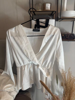 Vit omlottblus från Missay - Stilren vit omlottblus från Missay med knytband i midjan och v-ringad hals. Blusen har långa ärmar och ett lätt, flowy tyg som ger en avslappnad men chic look. Perfekt att matcha med jeans eller kjol för en trendig outfit.
