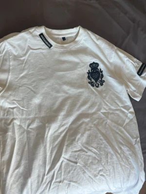 Vit t-shirt från Dolce & Gabbana - Snygg vit t-shirt från Dolce & Gabbana med svart broderad logga och emblem på bröstet med kristall liknande delar. Klassisk rund halsringning och korta ärmar. Tillverkad i mjuk bomull som känns skön mot huden. Perfekt för dig som gillar stilrena designer med exklusiv känsla.