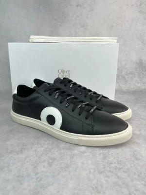 Svarta sneakers från Oliver Cabell - Stilrena svarta sneakers i läder från Oliver Cabell med vit sula och vit detalj på sidan. Skorna har svarta skosnören med text och vit hälflik. Perfekta för dig som gillar minimalistisk och modern design.
