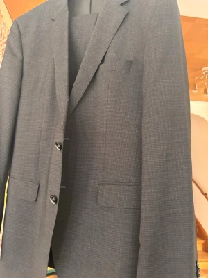 Mörkgrå kostym med stretch - Elegant mörkgrå kostym med ett diskret rutmönster som ger ett sofistikerat intryck. Kavajen är tvåknäppt och har klassiska slag, fickor med lock samt en praktisk innerficka. Stretchdetaljer bidrar till extra komfort och en smidig passform.  Ett utmärkt val för dig som söker en stilren bekväm outfit som fungerar lika bra till arbete som vid mer uppklädda tillfällen och eller student..