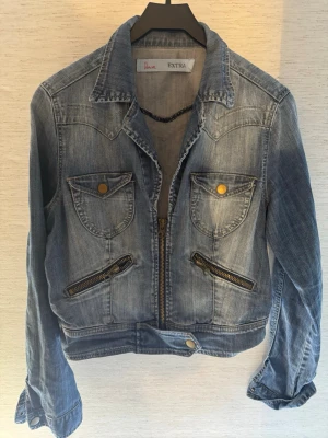 Blå jeansjacka från Extra - Snygg blå jeansjacka från Extra med klassisk krage, två bröstfickor med guldiga knappar och två snedställda dragkedjefickor. Jackan har en kortare passform och ett bälte med knapp nertill för extra detalj. Perfekt för en cool och avslappnad stil.