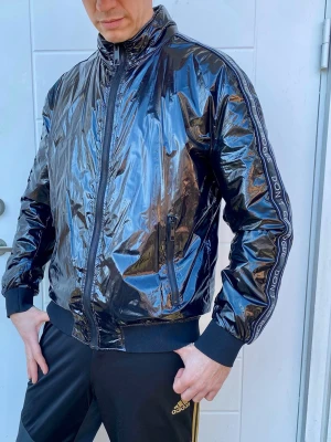 Svart glansig bomberjacka Zara - Ikonisk Svart glansig bomberjacka från Zara  med dragkedja framtill, ribbade muddar och krage. Jackan har logotypband längs ärmarna och två fickor med dragkedja. Materialet är syntetiskt och ger en cool, futuristisk look.