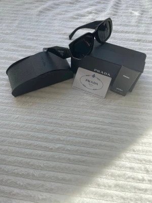 Prada Solglasögon  - Säljer ett par Prada solglasögon, de är helt svarta med silver logo på sidorna. Det som kommer med solglasögonen är originallådan, originalfodral, putsduk, certifikat och en manual. Säljer det för 900kr. Skickar paketet samma dag eller dagen efter ☺️