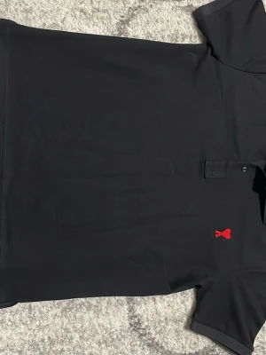 Svart pikétröja från Ami Paris - Klassisk svart pikétröja från Ami Paris med röd broderad logga i form av ett hjärta och bokstaven A på bröstet. Tröjan har korta ärmar, krage och knappar framtill. Perfekt till jeans eller kjol för en clean look.