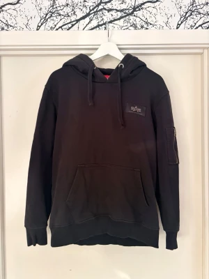 Svart hoodie från Alpha Industries - Svart hoodie från Alpha Industries med klassisk känguruficka, huva med dragsko och patch på bröstet. Ärmen har en cool ficka med dragkedja och extra detaljer. Perfekt för dig som gillar streetwear och vill ha en stilren look. Camoflage mönster. 950kr nypris mitt pris 400kr.