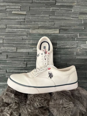 Vita sneakers från U.S. Polo Assn. - Storlek 42 - Snygga vita sneakers från U.S. Polo Assn. i canvasmaterial med lågt skaft. Skorna har vita skosnören, svarta och röda detaljer vid öglorna samt en broderad logga på sidan. Yttersulan är vit med en tunn svart rand runt om. Storlek 42. Aldrig använda. Säljer ett par likadana men i blått.