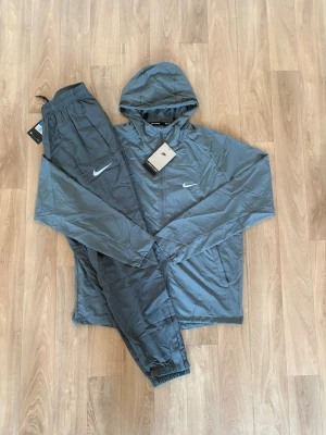 Grå Nike miler tracksuit - Snyggt grått träningsset från Nike med matchande jacka och byxor. Jackan har huva, dragkedja framtill och två fickor. Byxorna har resår i midjan och vid benslut. Perfekt för träning eller chill. Nike-logga på både jacka och byxa.