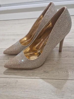 Glittriga roseguld pumps med strass - Säljer ett par snygga pumps i roseguld med spetsig tå och täckt av glittrande strass. Skorna har smal klack och insida i guldigt material som ger en lyxig känsla. Perfekta för dig som vill sticka ut och glänsa lite extra.