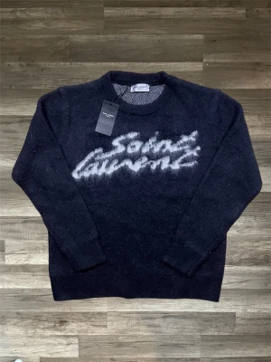 Svart Saint Laurent stickad tröja - Säljer en fluffig mörkgrå stickad tröja från Saint Laurent med vit text i grafisk stil över bröstet. Tröjan har rund halsringning och långa ärmar. Materialet ser ut att vara mohairblandning för extra mjuk känsla.