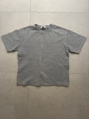 Grå t-shirt från H&M med struktur - En stilren grå t-shirt från H&M i storlek 146/152. T-shirten har en diskret rutstruktur i materialet och klassisk rund halsringning. Perfekt för en clean och avslappnad look.