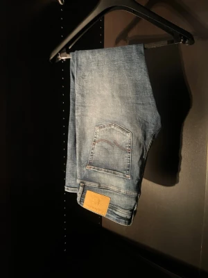 Blå slitna jeans från Jack & Jones - Säljer ett par blå jeans från Jack & Jones.  Jeans med slitningar och lätt tvättad look. Jeansen har klassisk femficksmodell, raka ben och detaljerad bakficka med Nudie-söm. Tillverkade i bomull för en bekväm känsla.