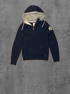 Mörkblå hoodie med dragkedja - Mörkblå hoodie med halv dragkedja framtill och vit insida på huvan. Stora vita bokstäver på ena ärmen och ljusgråa armbågslappar på baksidan. Ribbstickade muddar och mjukt material för extra komfort.