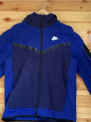 Blå Nike Tech hoodie - Snygg blå Nike Tech Hoodie med dragkedja. Hoodien har svarta detaljer och en stor huva. Knappt använd, ca 2-3 gånger.
