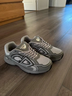 Dior b30 grå - Snygga grå sneakers från Dior med en chunky sula och coola detaljer i både mesh och läder. Skorna har tydliga Dior-loggor på sidan och tungan, samt matchande grå skosnören. Perfekta för dig som gillar streetwear och exklusiv design.