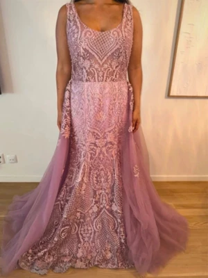 Ljusrosa långklänning med spetsdetaljer - Elegant långklänning med vackra spetsdetaljer och broderier. Klänningen har ärmlös design, djup v-ringning fram och öppen rygg. Kjolen är i flera lager tyll som ger en drömmig look. Perfekt för bal eller speciella tillfällen.