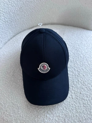 Svart keps från Moncler - Snygg svart keps från Moncler med klassisk böjd skärm och broderad Moncler-logga framtill. Justerbar rem baktill för perfekt passform. Tillverkad i slitstarkt tyg som ger en clean och stilren look.