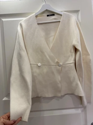 Vit omlottkofta från Gina Tricot - Snygg vit omlottkofta från Gina Tricot med v-ringning och två dekorativa knappar framtill. Koftan har lång ärm och är tillverkad i ett mjukt, stickat material.