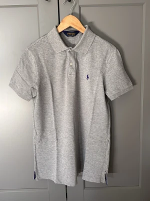 Grå polotopp från Polo Ralph Lauren - Klassisk grå polotopp från Polo Ralph Lauren med korta ärmar och krage. Toppens material är mjuk bomull och den har två knappar framtill samt en broderad logga i blått på bröstet. Perfekt för en avslappnad och stilren look.