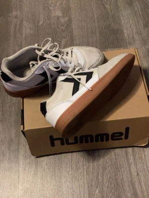 Vita Hummel sneakers med gummisula - Snygga vita sneakers från Hummel med klassiska svarta pilar på sidorna och brun gummisula. Skorna har en mix av mocka och textilmaterial, samt vita snören. Perfekta för en sportig och avslappnad stil.