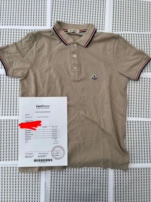 Beige pikétröja från Moncler - Snygg beige pikétröja från Moncler med klassisk krage och knäppning framtill. Tröjan har Moncler-logga på bröstet och detaljer i rött, vitt och svart på krage och ärmslut. Tillverkad i mjuk bomull som känns skön mot huden.