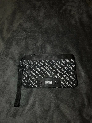 Svart kuvertväska Versace Jeans Couture - Snygg svart kuvertväska från Versace Jeans Couture med vitt logomönster över hela väskan. Väskan har en avtagbar handledsrem och dragkedja upptill. Tillverkad i slitstarkt tygmaterial och har dekorativa nitar framtill.