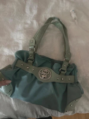 Grön handväska med silverdetaljer - Snygg grön handväska med dekorativa silverdetaljer och ett stort emblem framtill. Väskan har dubbla handtag i matchande färg och nitar längs kanterna. Insidan är rymlig med ljus textilfoder och tryckknappsstängning. Perfekt för dig som gillar statement-accessoarer.