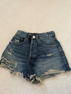 Högmidjade blå jeansshorts med slitningar - Säljer ett par högmidjade blå jeansshorts med råa kanter och slitna detaljer framtill. Klassisk femficksmodell med knapp och dragkedja. Perfekta för sommaren och ger en avslappnad, trendig look.